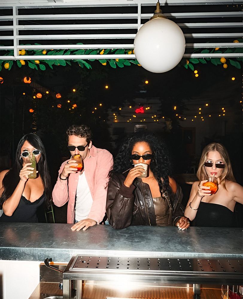 Four friends in sunglasses drinking at a bar under festoon lights