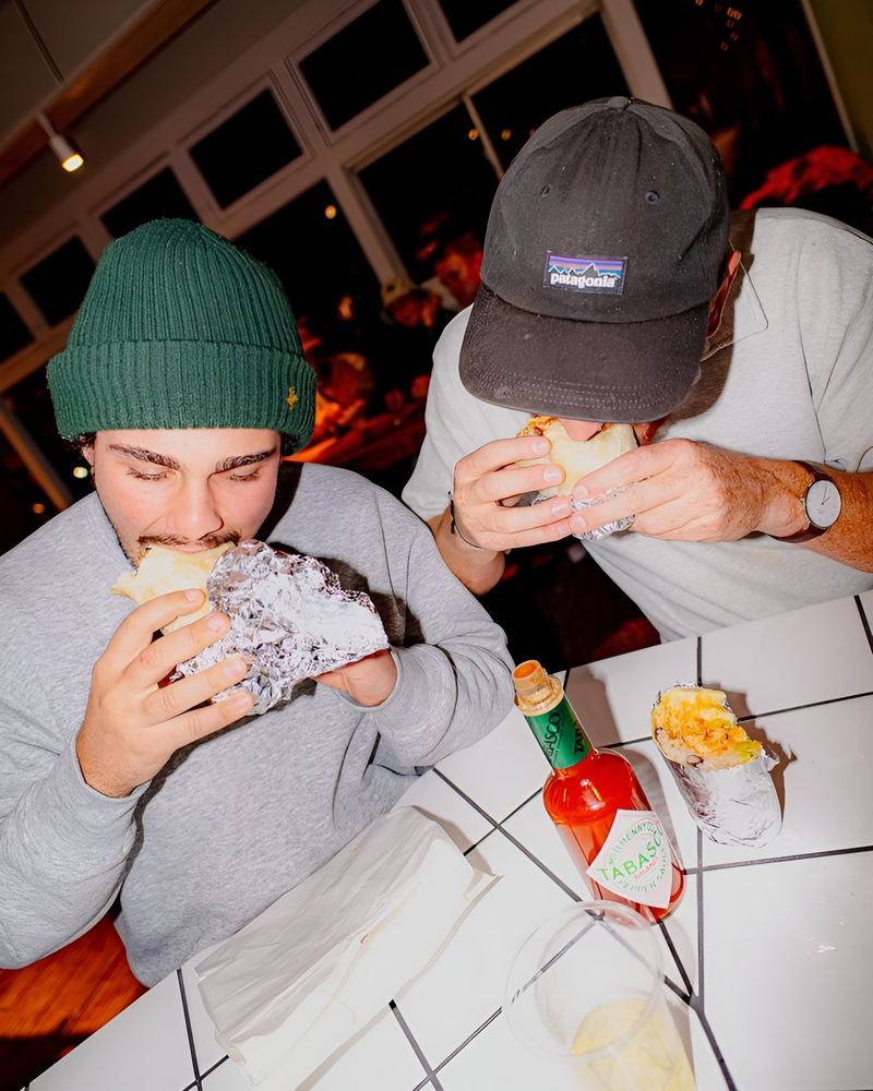 Two friends eating foil-wrapped burritos at a tiled table
