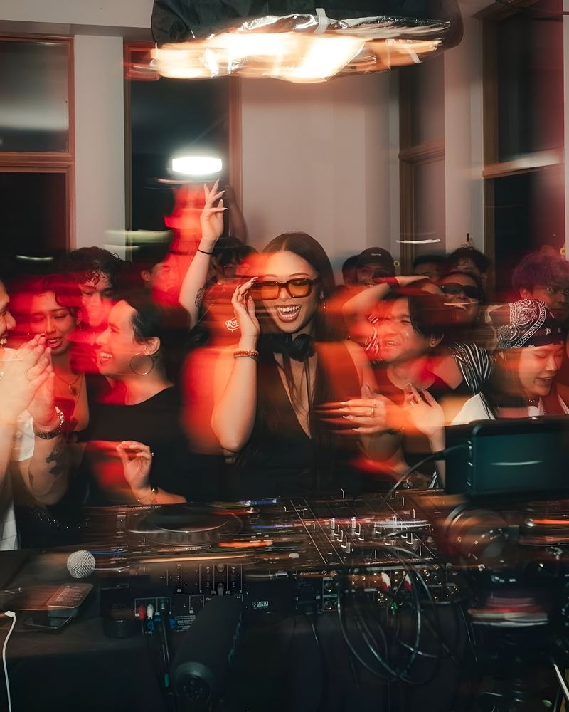 A DJ laughing behind the decks with a crowd around her