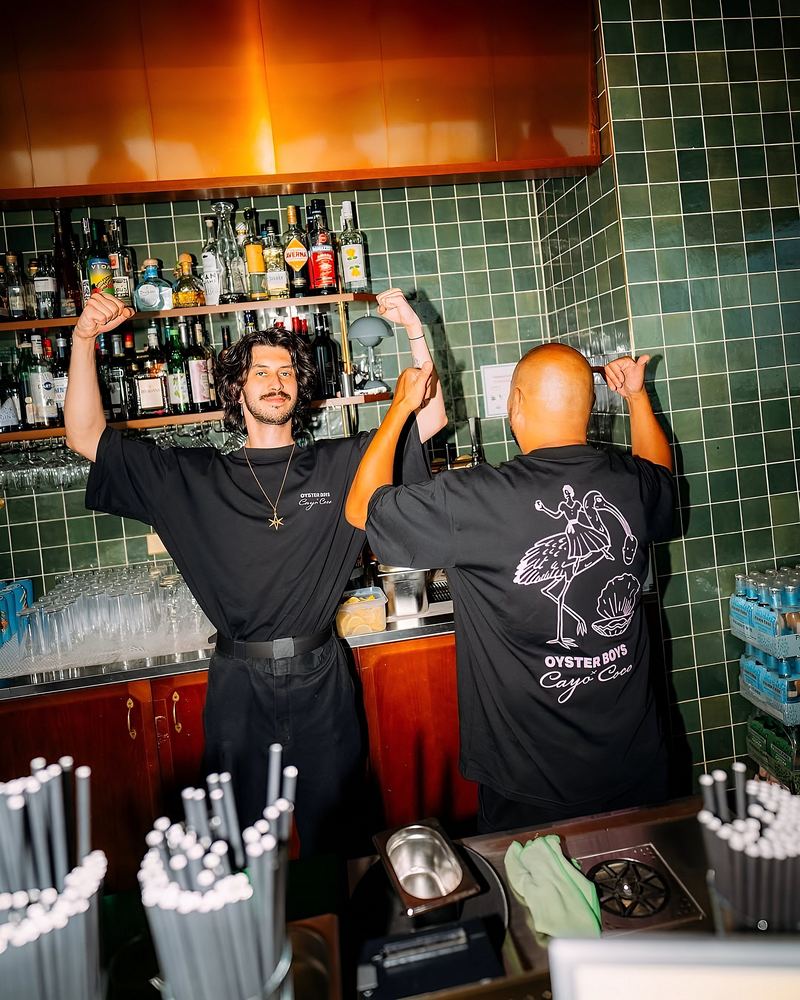 Two bartenders with arms raised behind a green tiled bar